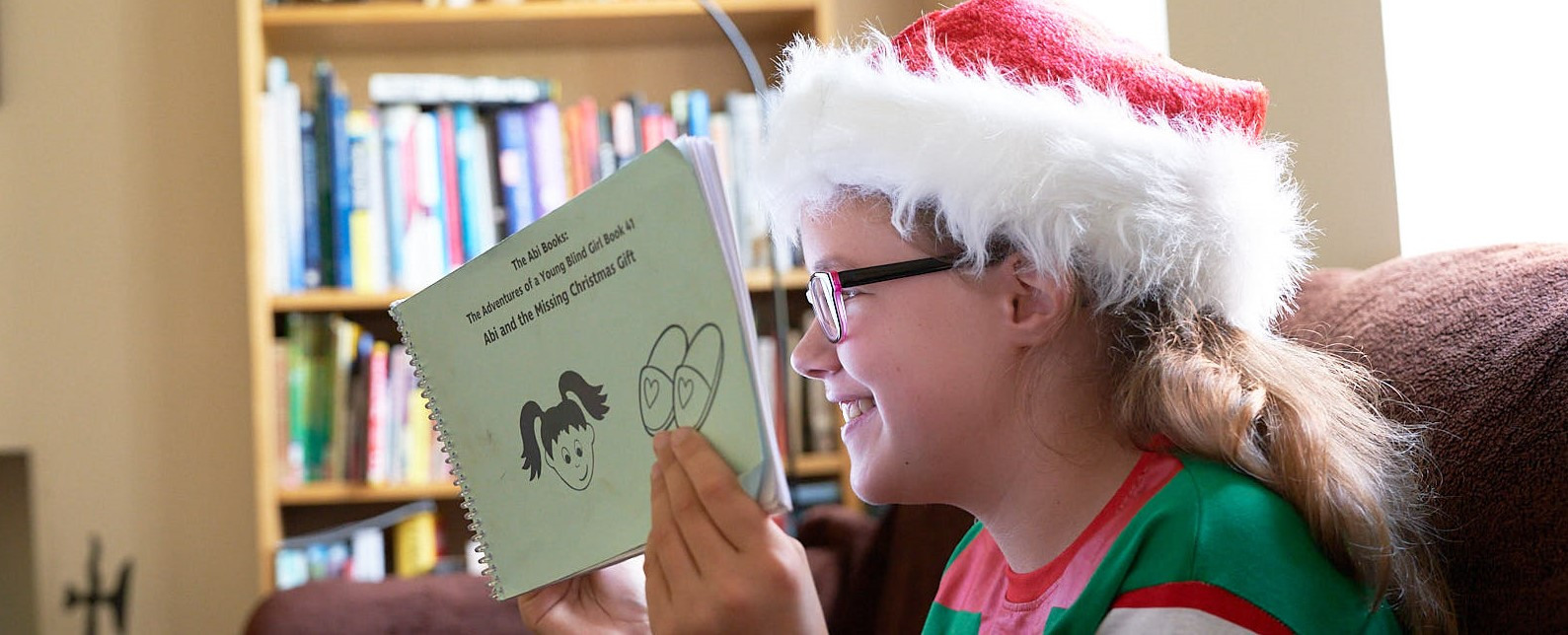 Smiling girl from the side wearing a Santa hat holding up a book reading it.