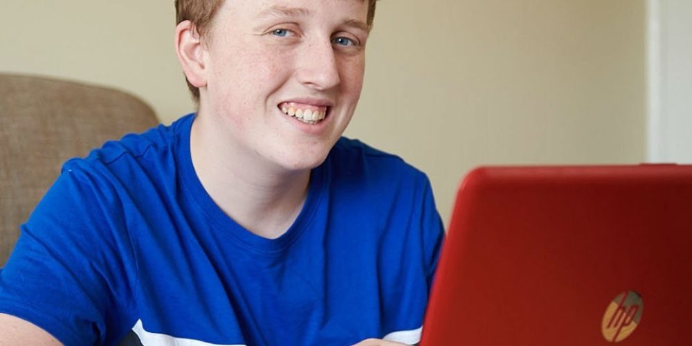 A smiling teenage boy sat at a desk with a open laptop