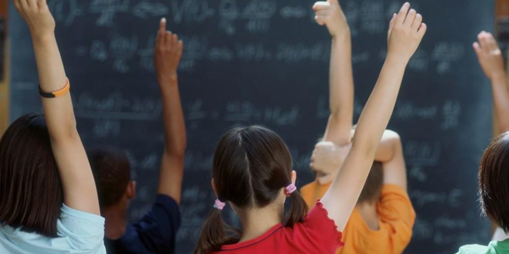 view from back of a classroom with childrens hands up to answer questions, blackboard in background