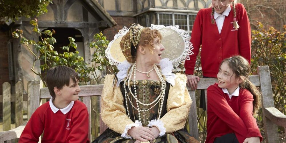 Queen Elizabeth the first outside a tudor style house with 3 modern school children