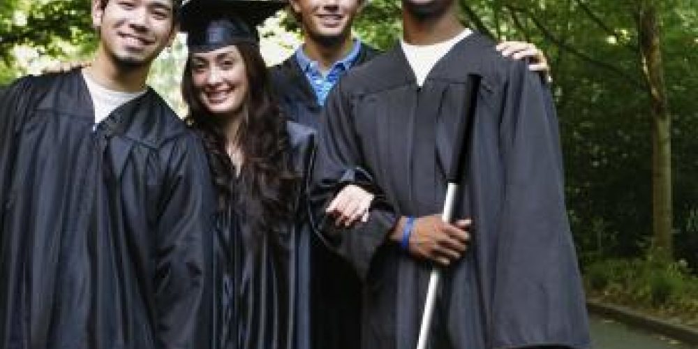 Four smiling students in graduation gowns and caps. The student on the far right is holding a long cane.