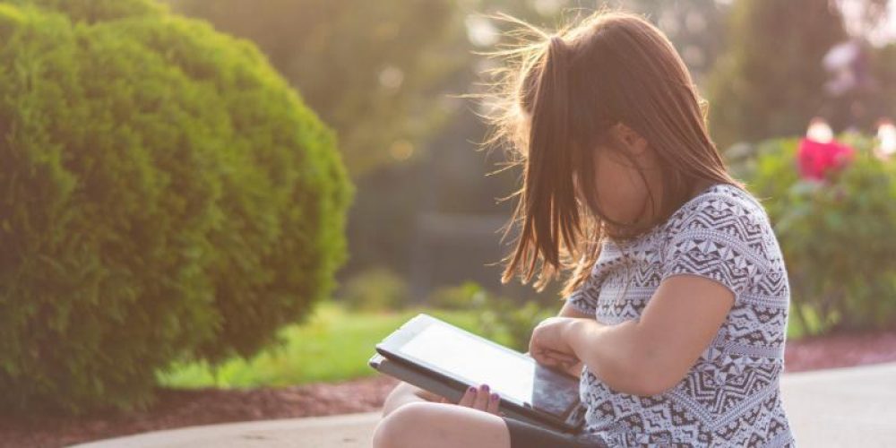 young girl sat outside in the sunshine looking down at a tablet she is holding