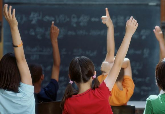 view from back of a classroom with childrens hands up to answer questions, blackboard in background