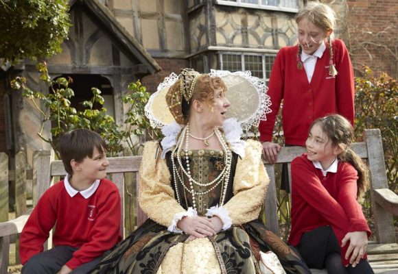 Queen Elizabeth the first outside a tudor style house with 3 modern school children