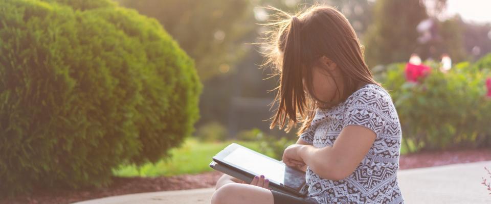 young girl sat outside in a sunny garden reading on a tablet